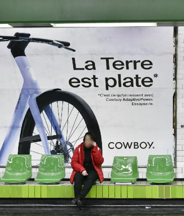 A person in a red jacket sits on green chairs in a subway station, in front of an advertisement featuring a bicycle and the French text La Terre est plate* on a white background.