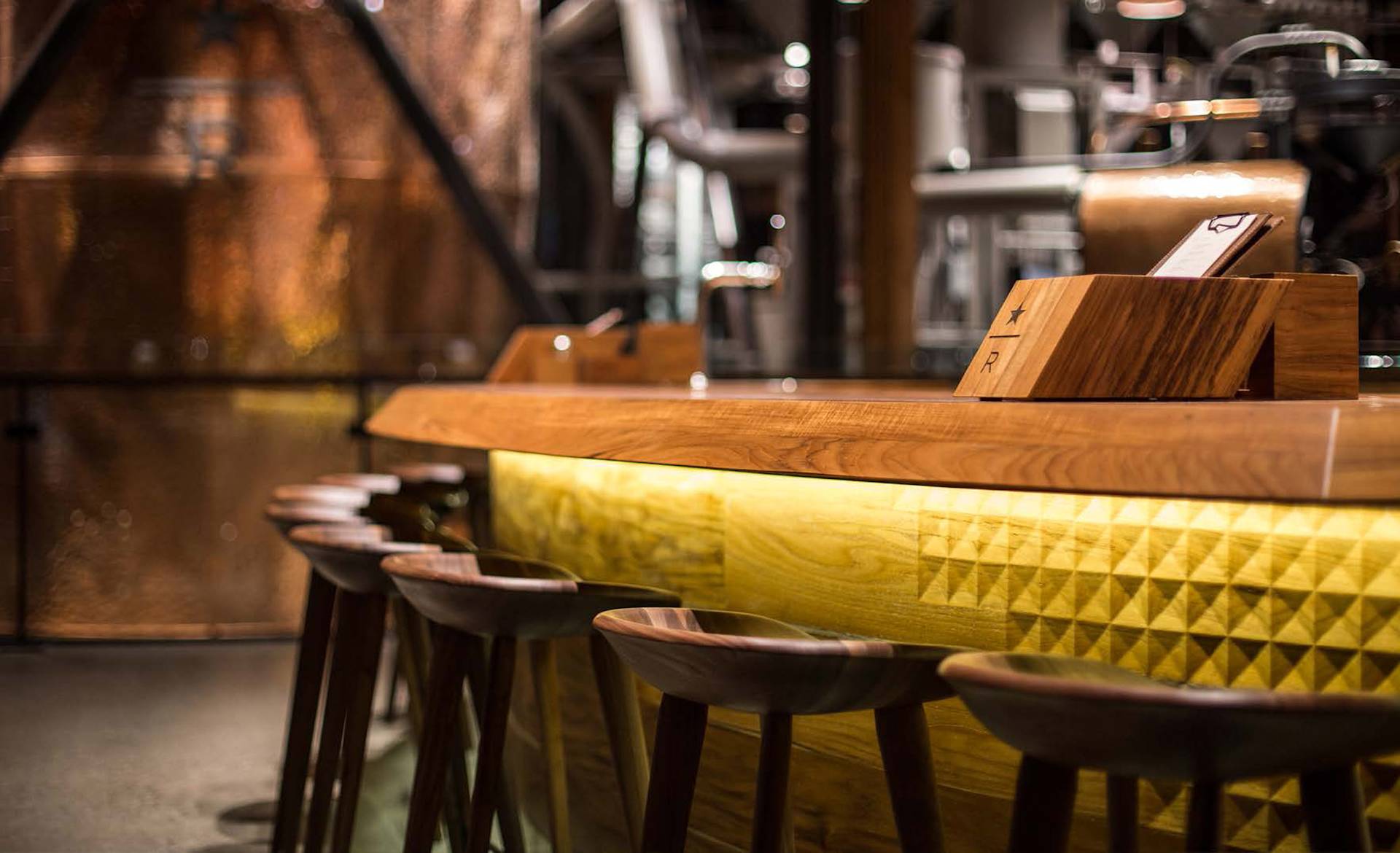 A modern bar with wooden stools lined up along a glowing wooden counter, featuring geometric patterns and warm lighting. Industrial-style equipment and copper tanks are visible in the blurred background.