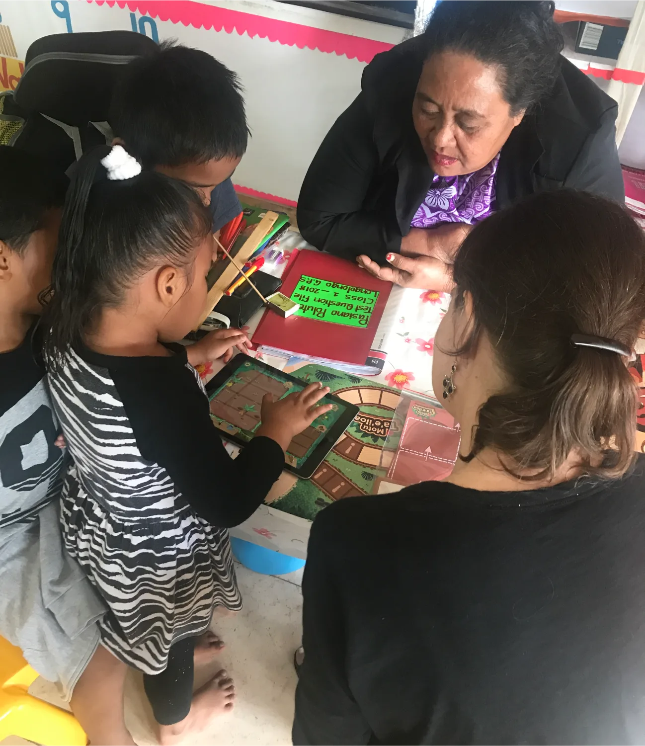 A group of young children and two adults are gathered around a table, engaging with colorful educational materials. One adult is speaking, while the children interact with books and cards.