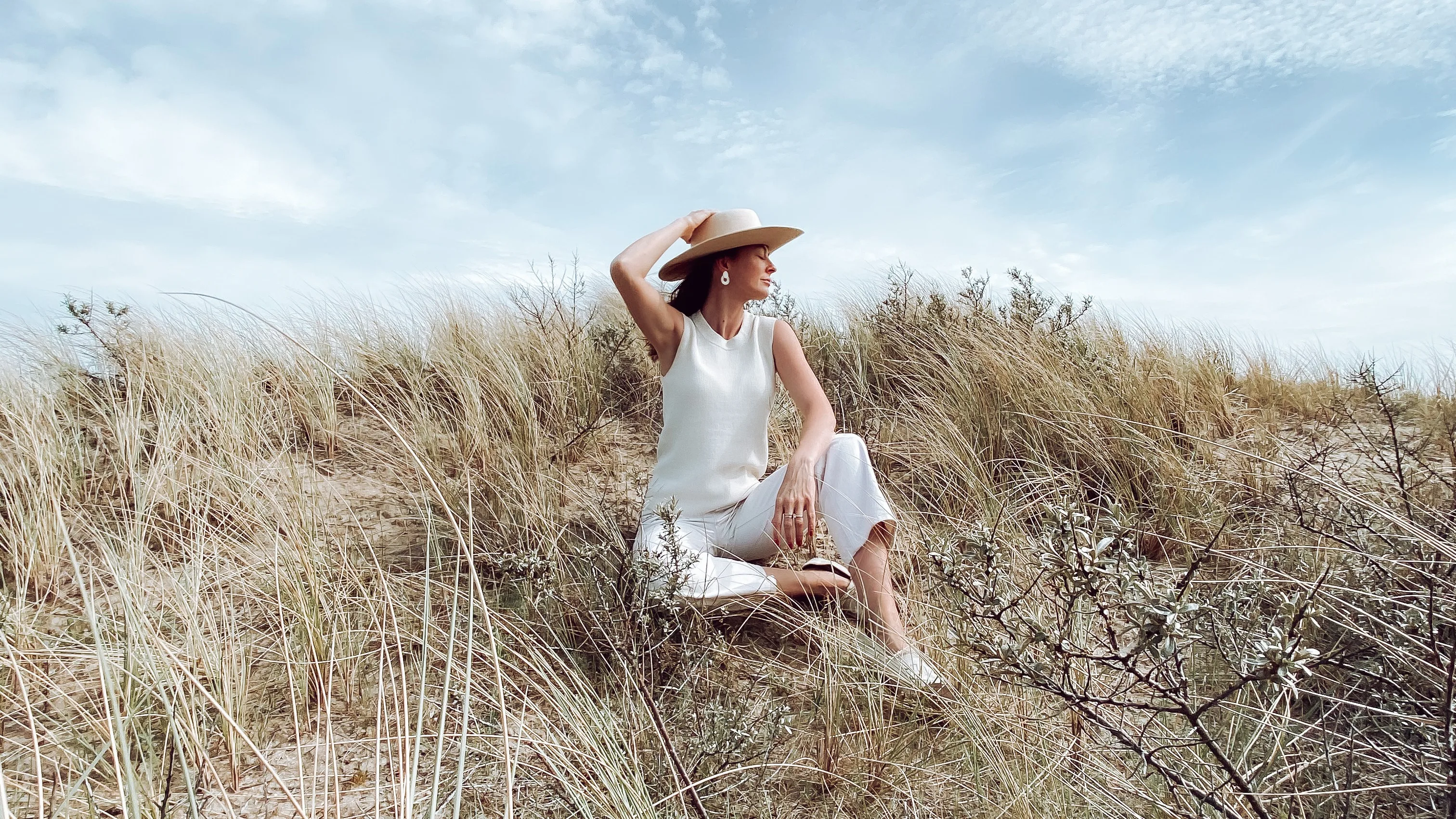 A woman in a white outfit and wide-brimmed hat sits on sandy grass dunes, looking to the side and smiling, with a blue sky and wispy clouds in the background.