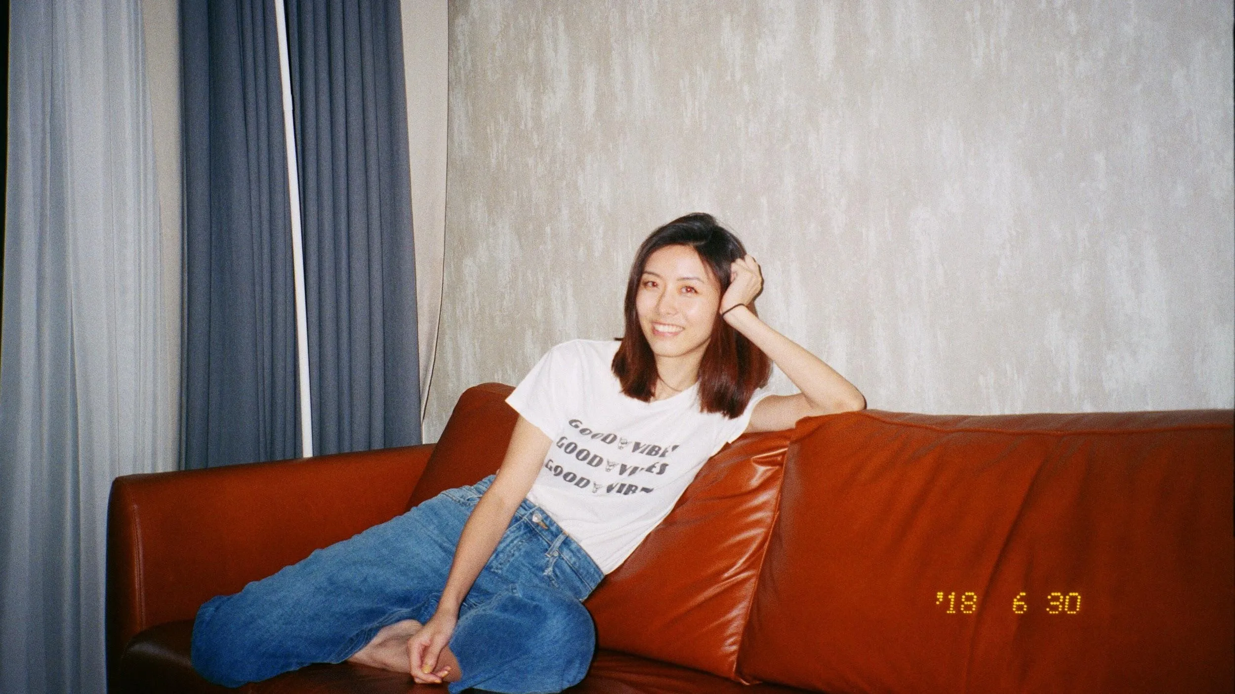 A woman with shoulder-length hair, wearing a white t-shirt and blue jeans, sits relaxed on a brown leather sofa, smiling at the camera. The date 18 6 30 is marked in yellow at the bottom right.