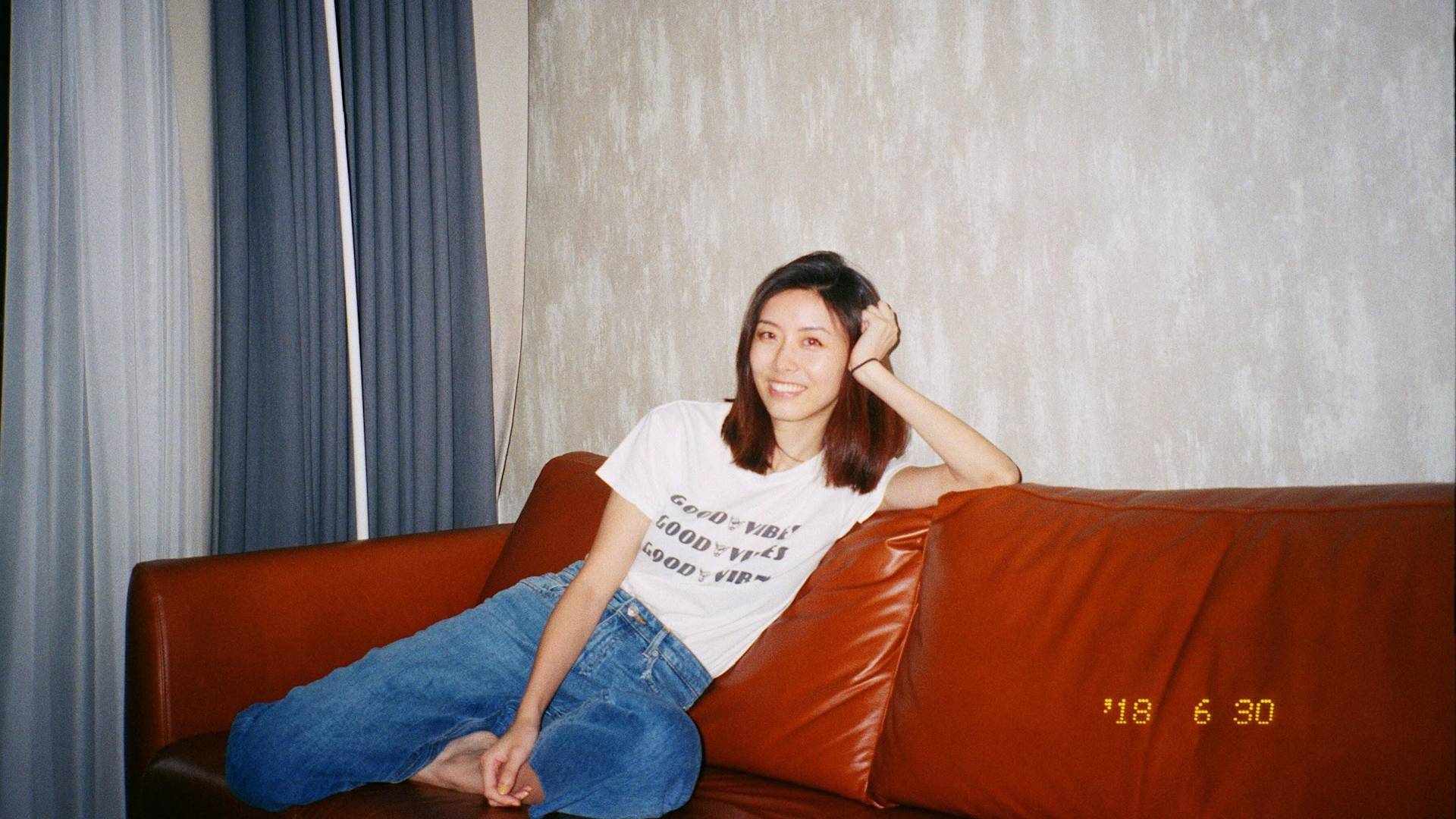 A woman with shoulder-length hair, wearing a white t-shirt and blue jeans, sits relaxed on a brown leather sofa, smiling at the camera. The date 18 6 30 is marked in yellow at the bottom right.