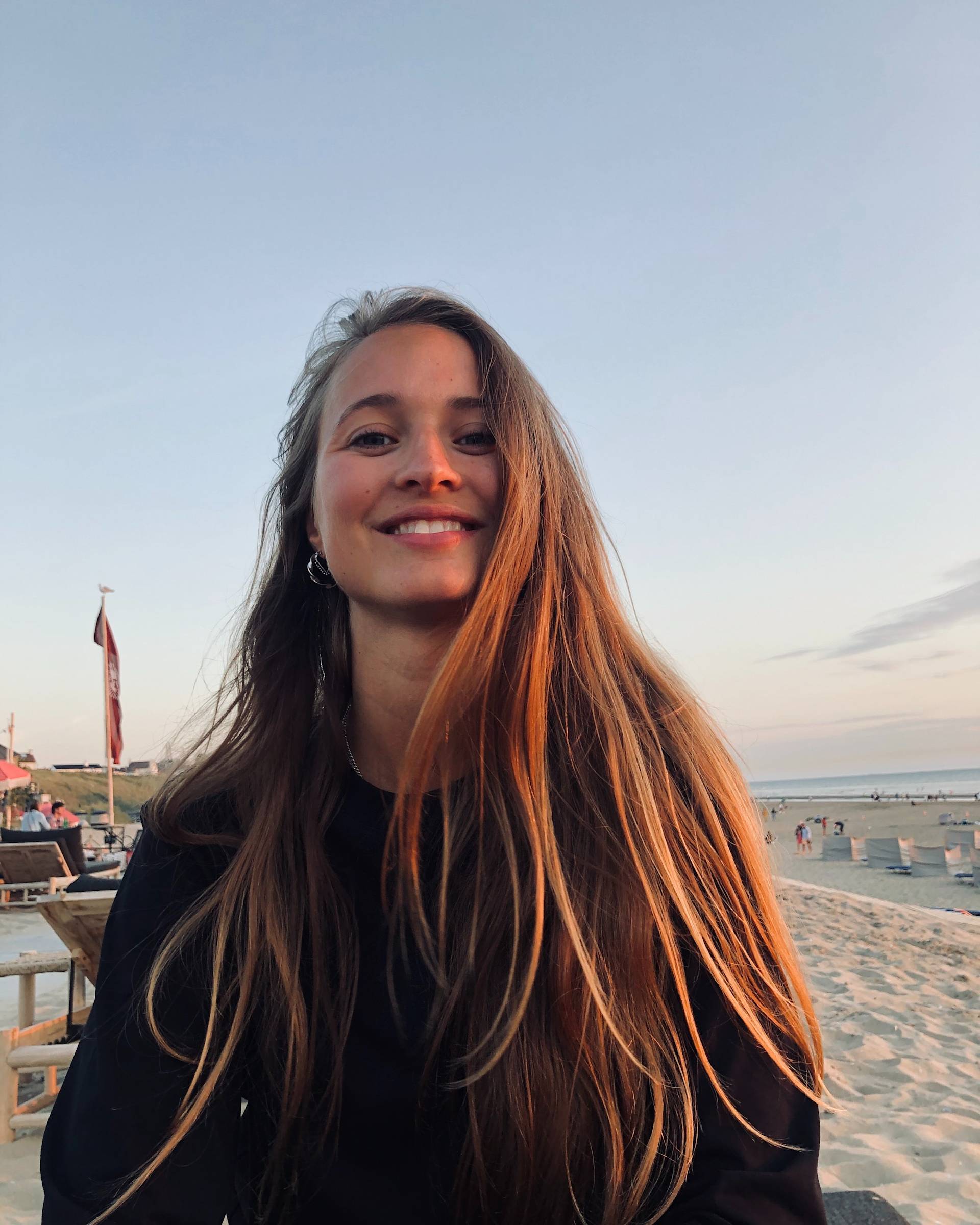 A young woman with long brown hair smiles at the camera while sitting on a beach in the evening, with the ocean and people in the background under a clear sky.