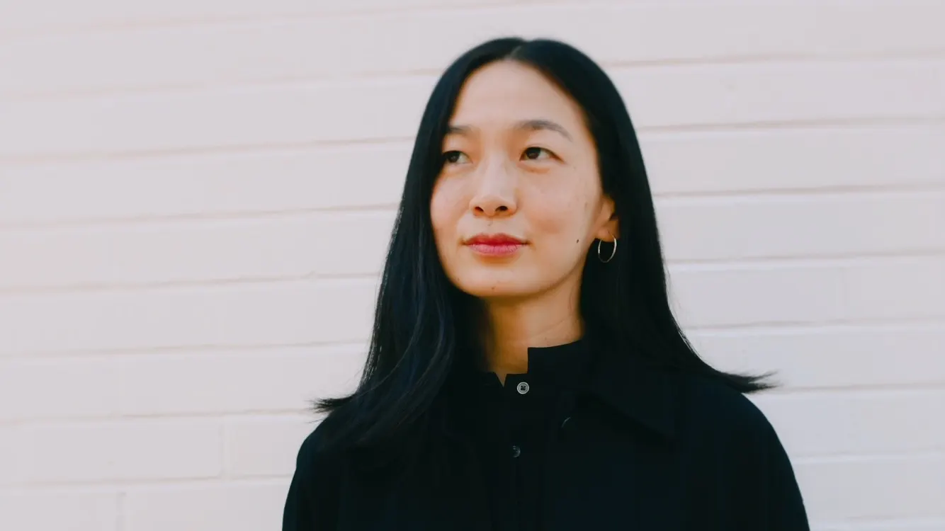 A woman with long black hair and hoop earrings stands against a white brick wall, wearing a black collared shirt and looking slightly to the side with a neutral expression.
