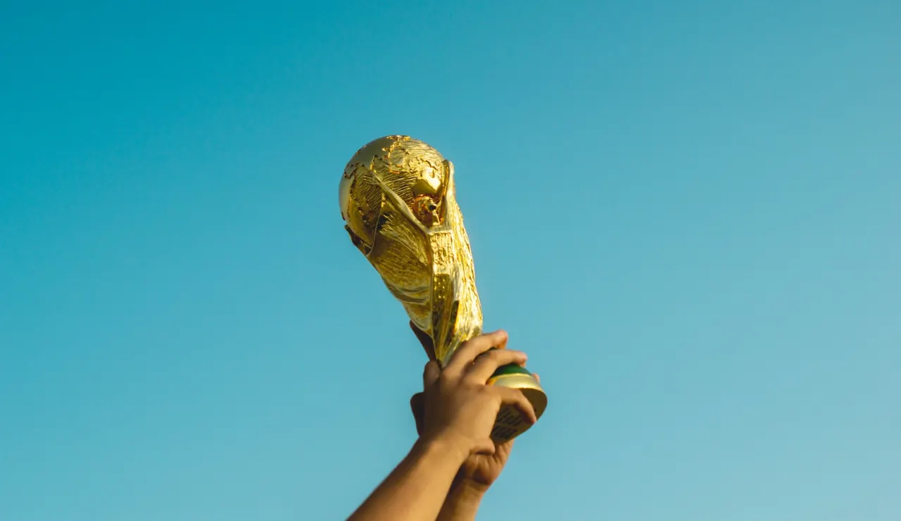 A person raises a golden trophy, resembling the FIFA World Cup, against a clear blue sky.