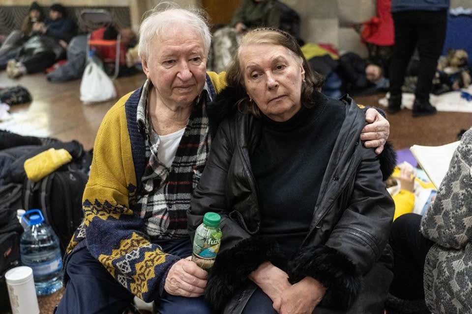 Two elderly people sit close together on the floor in a crowded indoor space, looking somber. One has an arm around the other, who holds a drink bottle. Belongings and other people are scattered in the background.