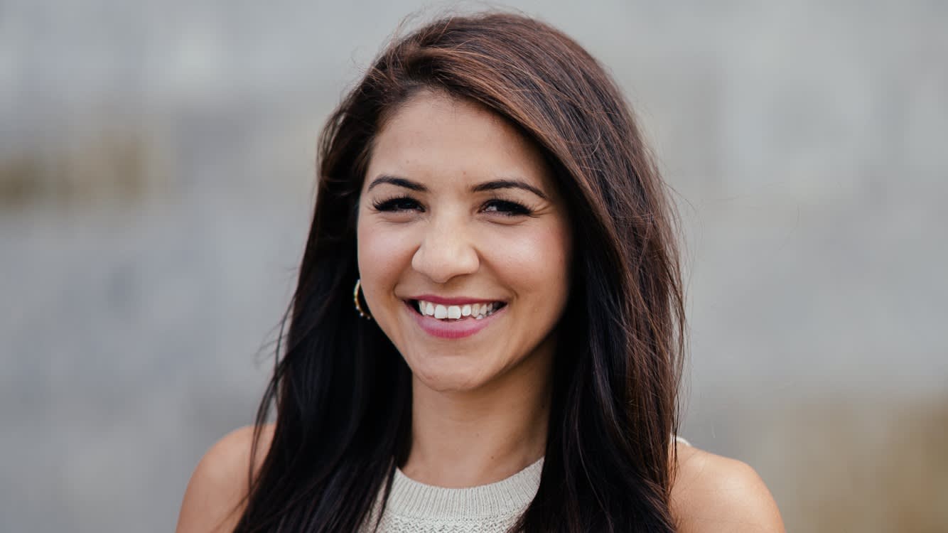 A woman with long dark hair smiles warmly at the camera, standing outdoors with a blurred gray background. She is wearing a light-colored sleeveless top and gold hoop earrings.