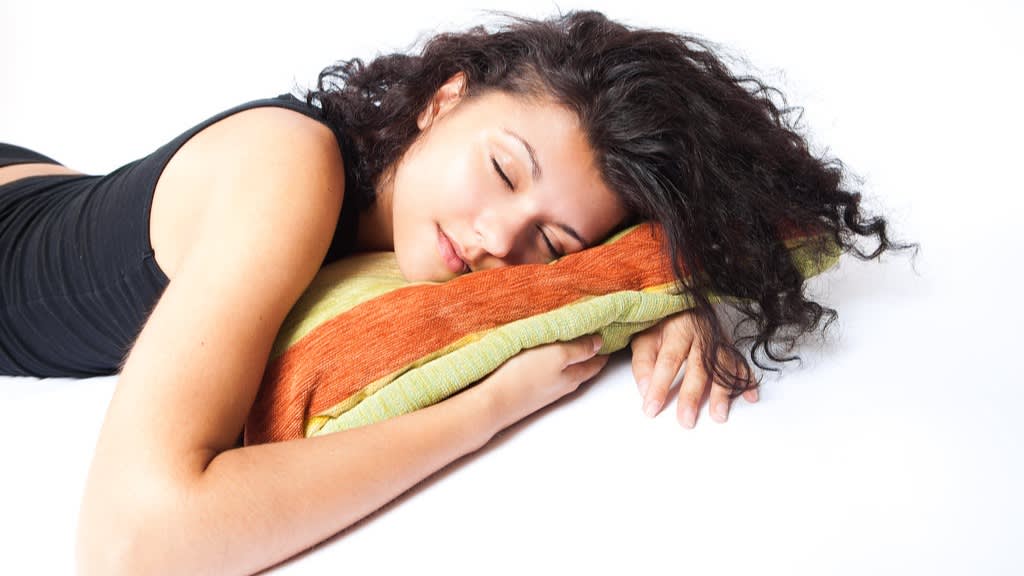 A young woman with curly hair sleeps peacefully on her side, resting her head on an orange and yellow pillow against a white background.