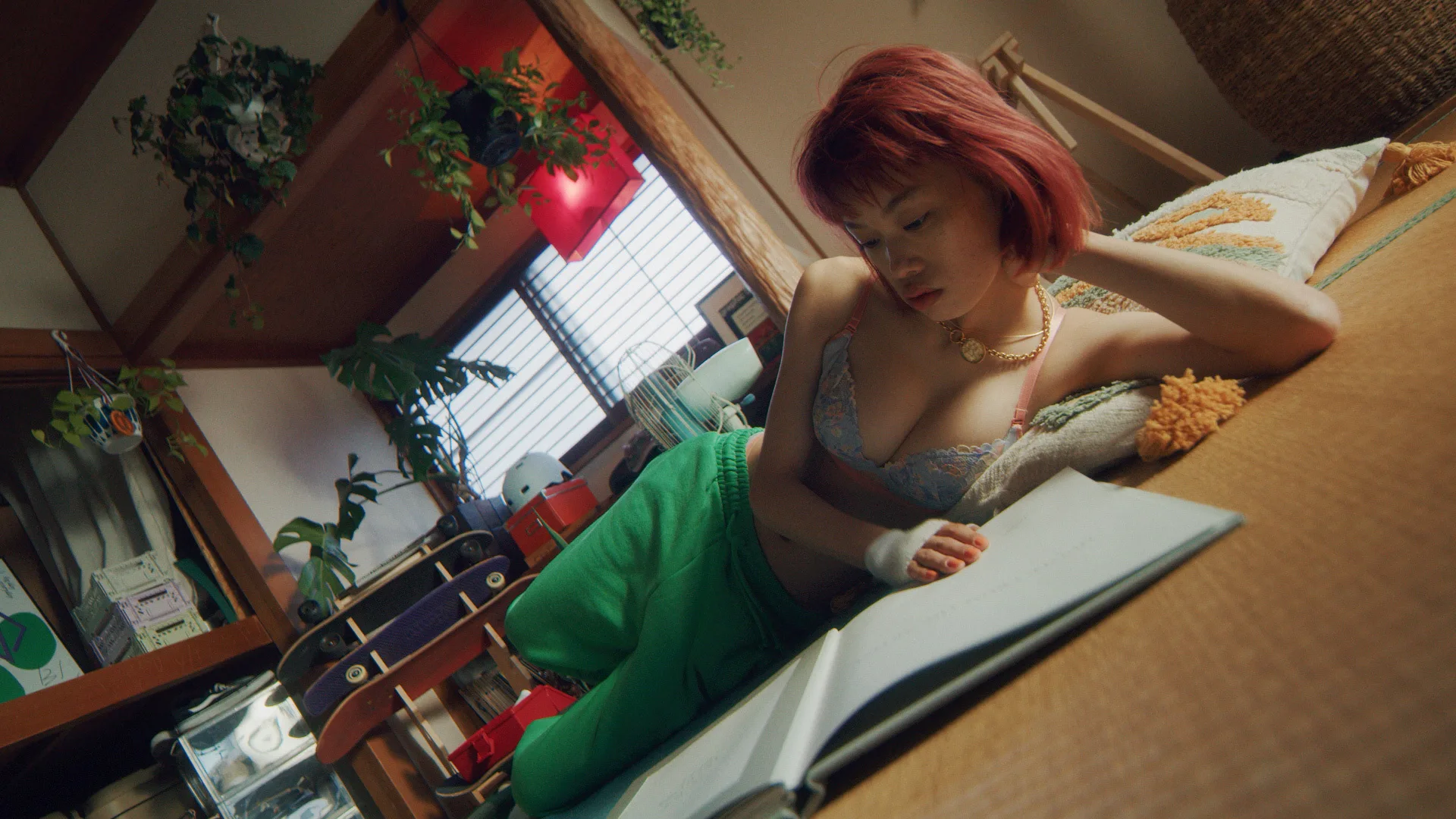 A young woman with short red hair lounges on the floor in a casual room, wearing a bralette and green pants, reading or writing in an open notebook. Houseplants hang above her and sunlight filters through a nearby window.