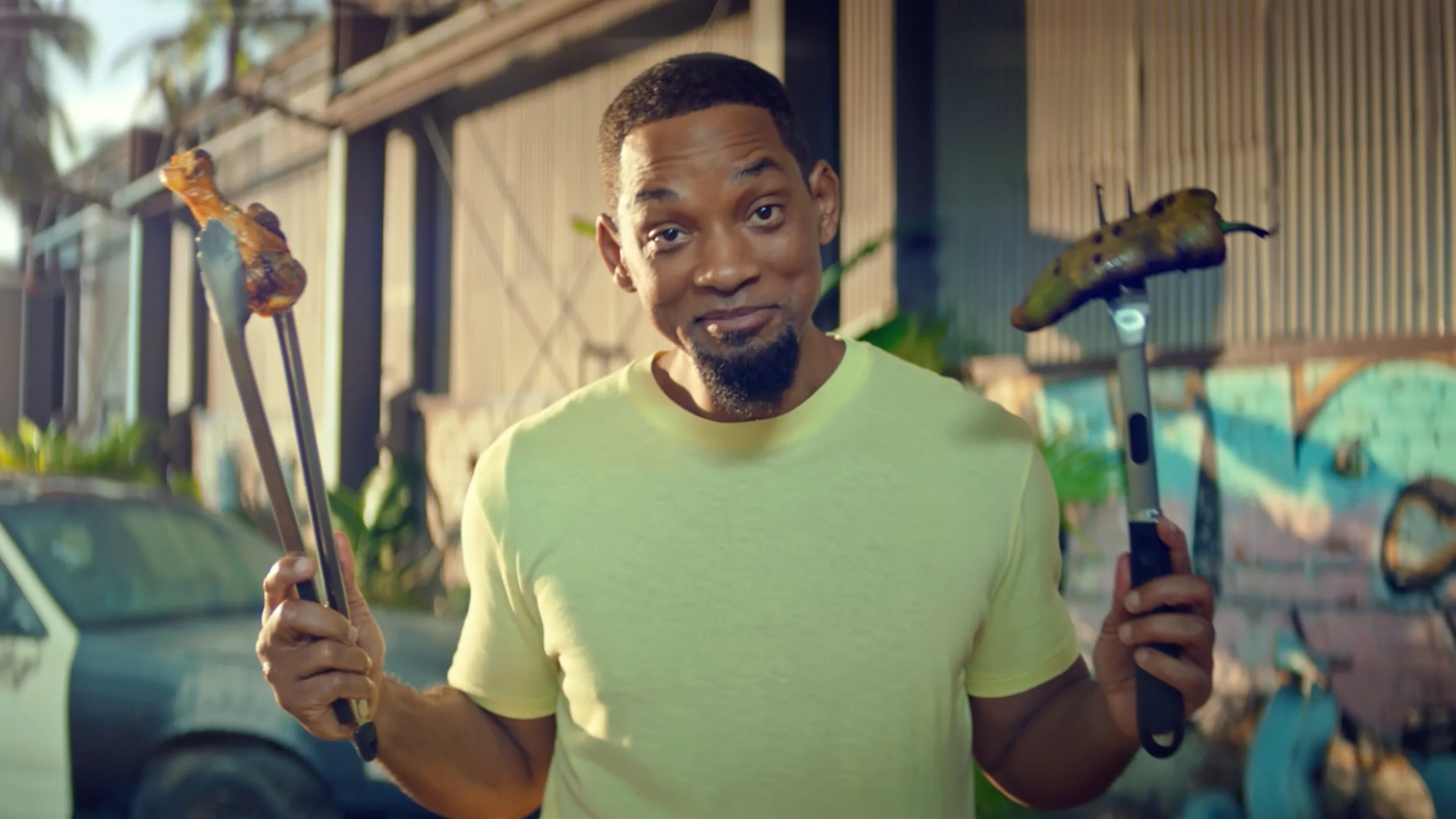 A man in a light green shirt stands outside, smiling and holding grilled food with tongs in one hand and a grilling fork in the other, with a building and greenery in the background.