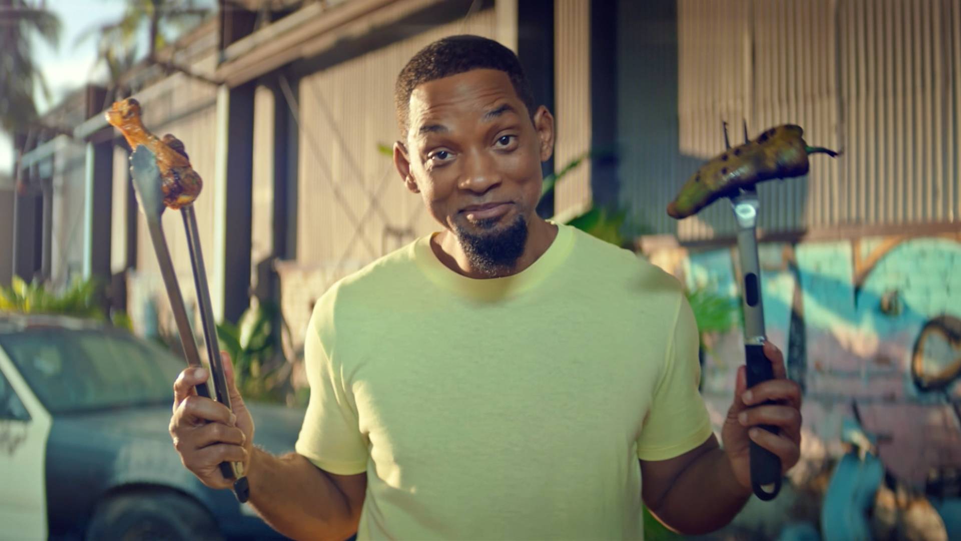 Will Smith in a light green shirt stands outside, smiling and holding grilled food with tongs in one hand and a grilling fork in the other, with a building and greenery in the background.