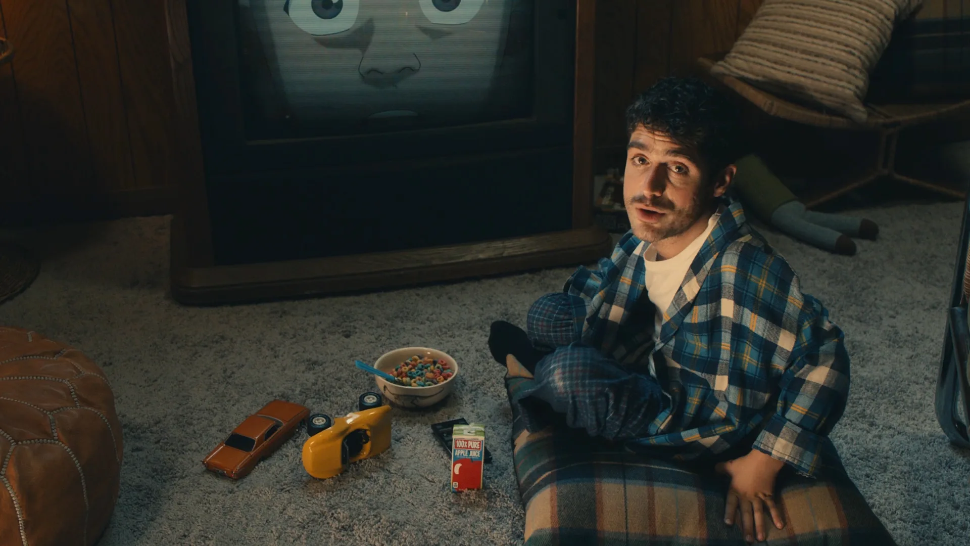 A young man in plaid pajamas sits on the floor with a pillow, surrounded by toy cars, cereal, and juice. Behind him, an old TV shows a cartoon face with wide eyes. The room has carpet and wood-paneled walls.
