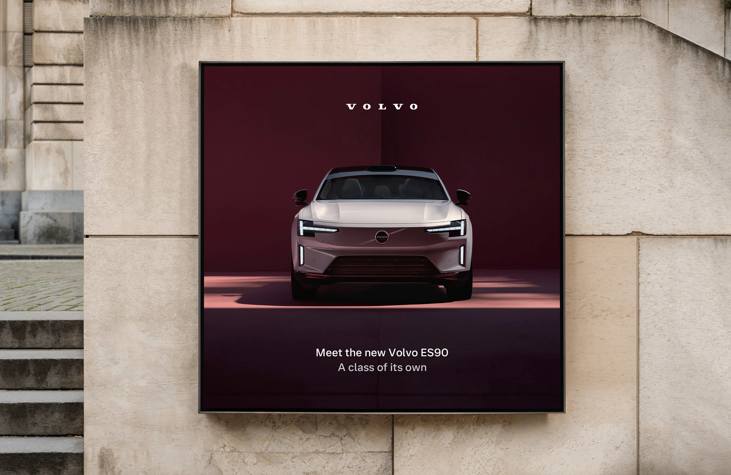 A poster on a stone wall displays the front view of a silver Volvo ES90 with vertical headlights, the Volvo logo above, and the text: Meet the new Volvo ES90. A class of its own.