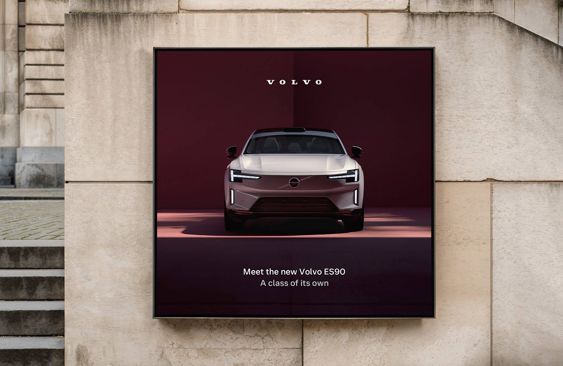 A poster on a stone wall displays the front view of a silver Volvo ES90 with vertical headlights, the Volvo logo above, and the text: Meet the new Volvo ES90. A class of its own.