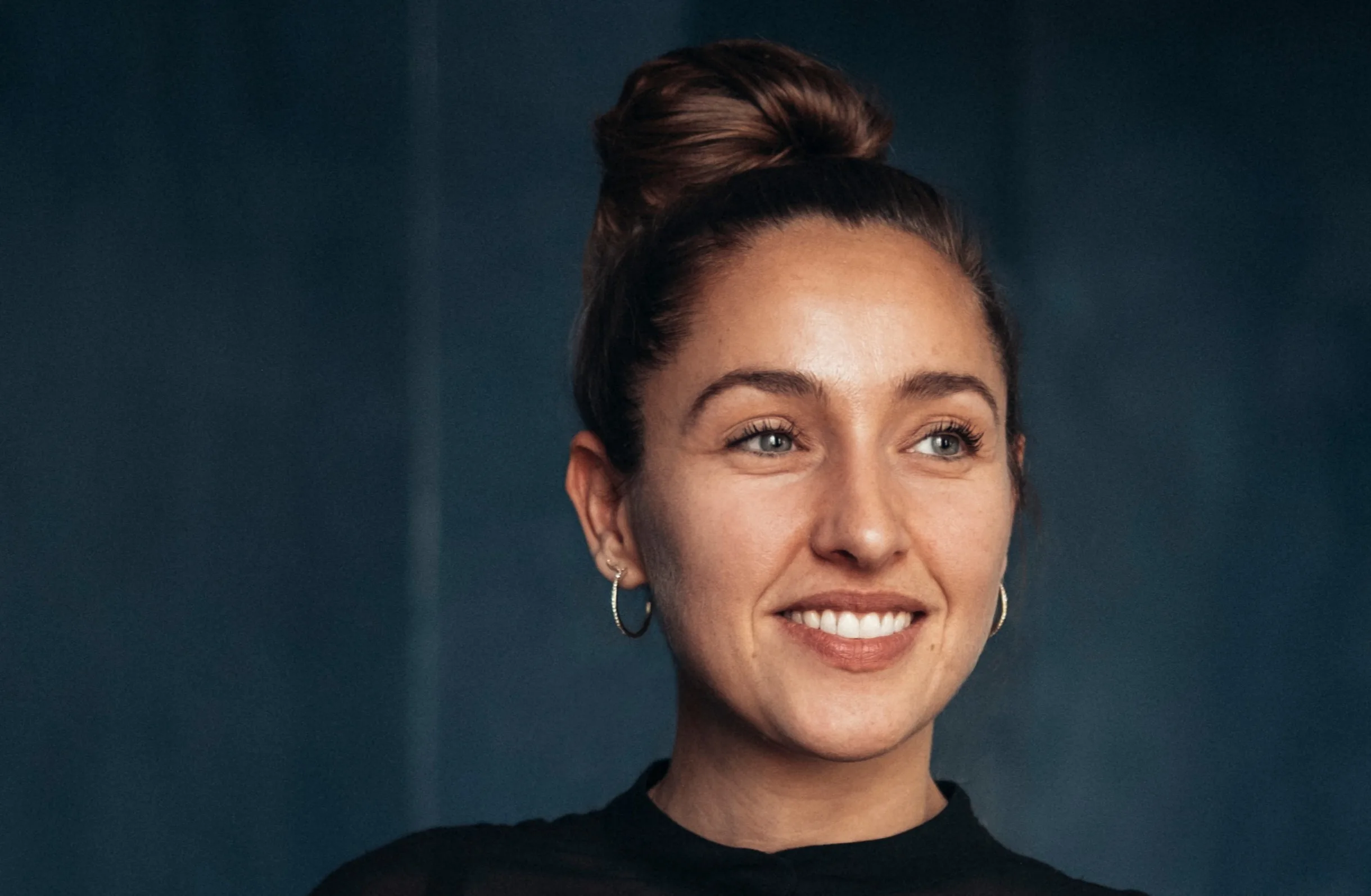 A woman with light skin and brown hair in a bun, wearing hoop earrings and a black top, smiles while looking slightly to the side against a dark, blurred background.