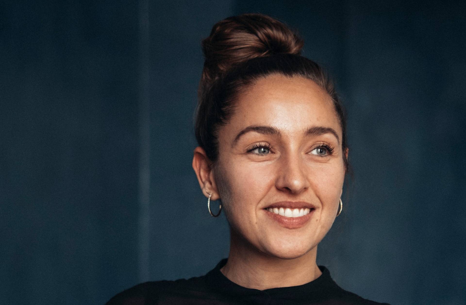 A woman with light skin and brown hair in a bun, wearing hoop earrings and a black top, smiles while looking slightly to the side against a dark, blurred background.