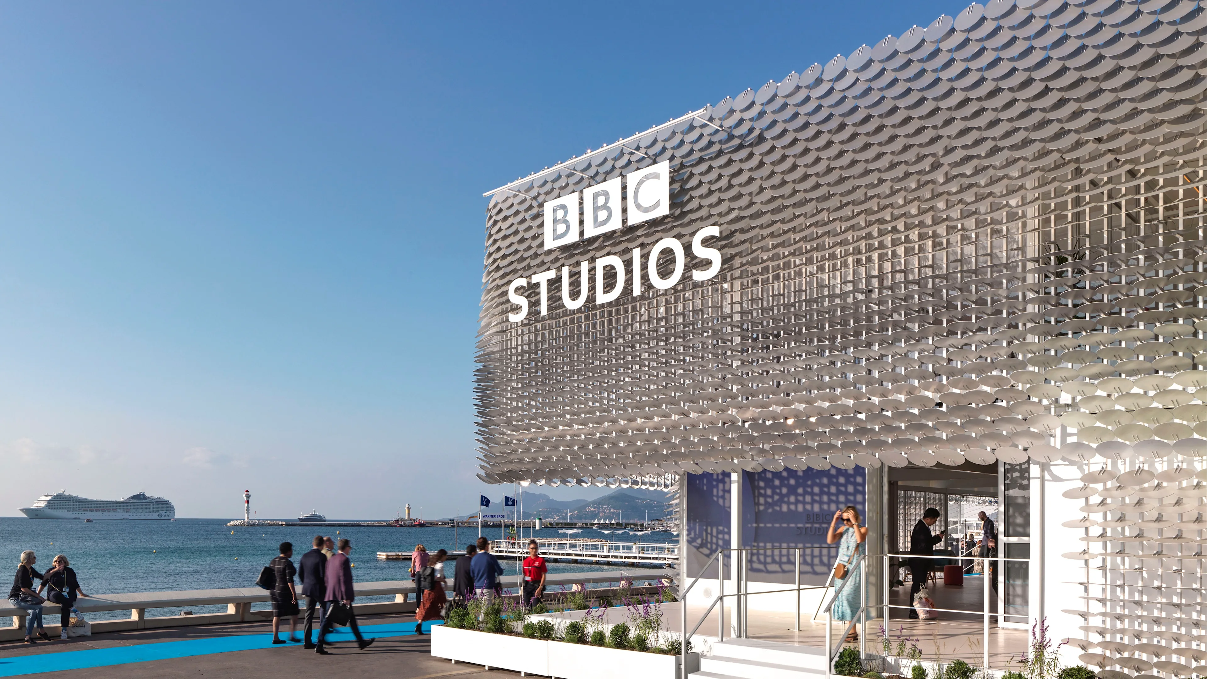 A modern BBC Studios building with a unique reflective facade sits by the sea, people walking nearby on a sunny day, and a cruise ship visible in the background.
