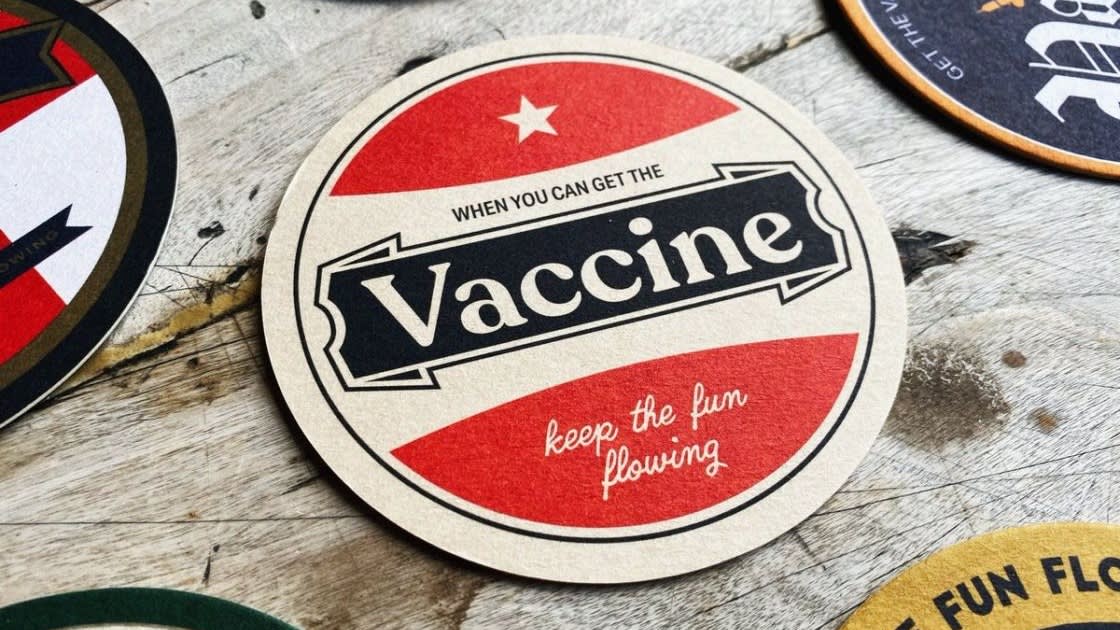 A round coaster with a red and white design reads When you can get the Vaccine, keep the fun flowing in bold and script fonts, placed on a wooden surface among other coasters.