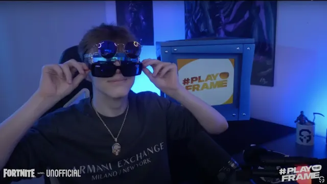 A person wearing multiple pairs of sunglasses smiles in front of a blue box with a #PLAYTHEFRAME sign. The room is lit with blue lighting, and the Fortnite and #PLAYTHEFRAME logos appear on the screen.