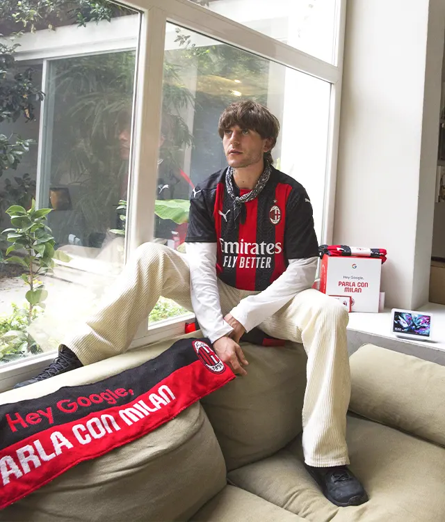 A young man wearing an AC Milan jersey and bandana sits on a sofa indoors, holding a red AC Milan scarf. A window with greenery outside is behind him, and AC Milan merchandise is on a nearby table.