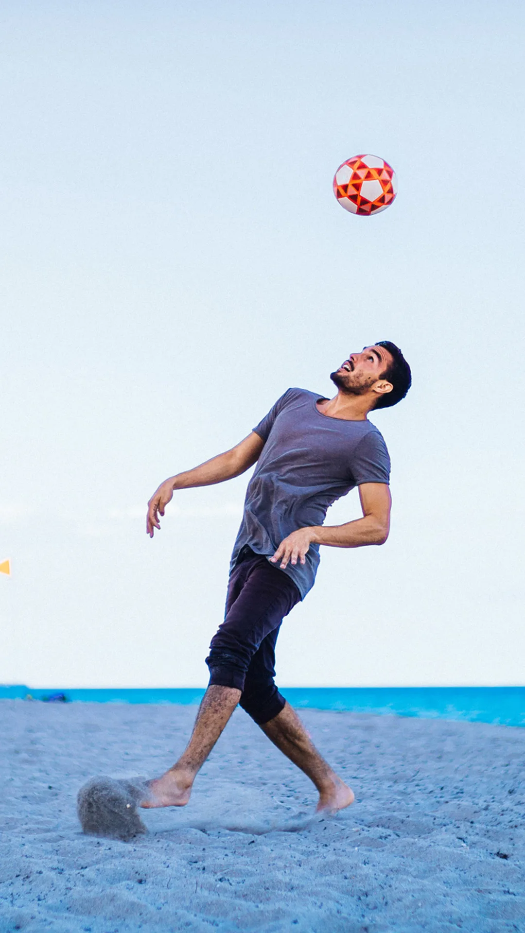 A man playing soccer on the beach, looking up and preparing to head a red and white ball in mid-air, with the ocean and blue sky in the background.