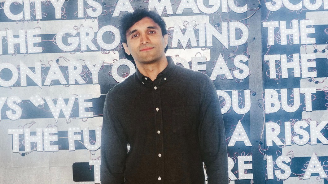 A man with short dark hair is standing indoors in front of a neon-lit sign with large white letters. He is wearing a dark button-up shirt and is looking at the camera with a neutral expression.