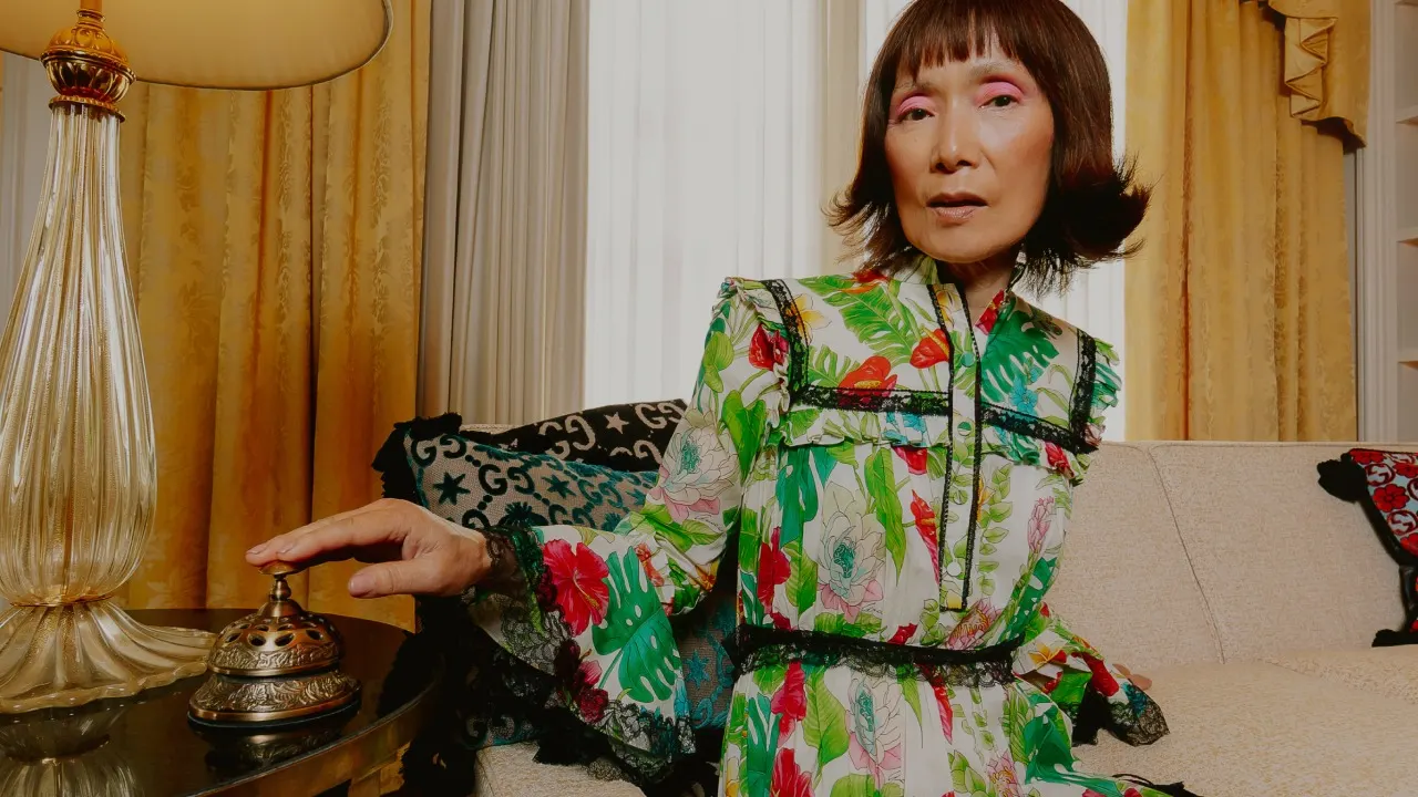 A woman with short hair and bold pink eye makeup, wearing a floral dress, sits on a cream sofa in a warmly lit room with yellow curtains. She is reaching out to press a small decorative bell on a side table.