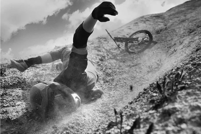 A person wearing a helmet and protective gear falls on a rocky slope while their bicycle lies behind them, partially airborne. Dust and debris are scattered, suggesting a recent crash.