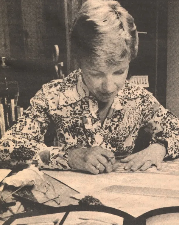 A person with short hair and a patterned shirt sits at a desk, writing or drawing on paper, focused on their work. Various papers and objects are scattered on the desk. The image is in black and white.