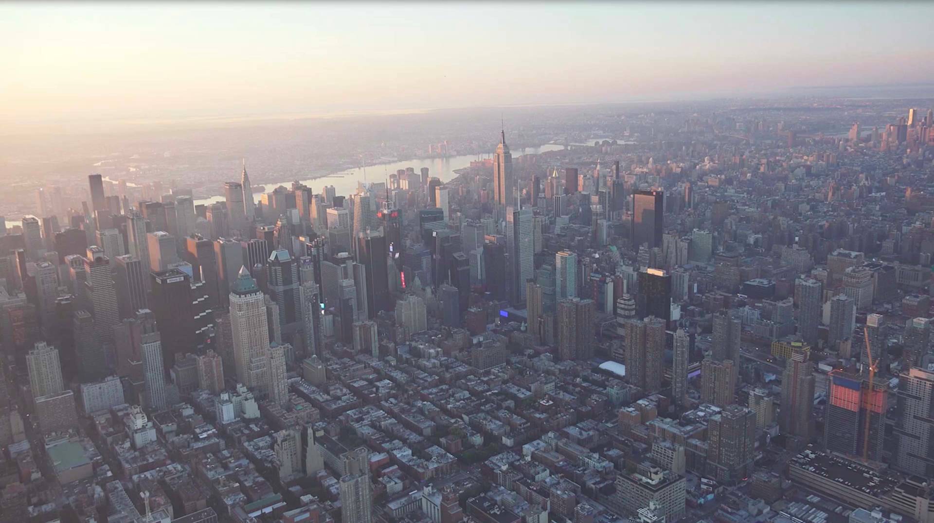 Aerial view of New York City at sunrise, showing densely packed skyscrapers, the Empire State Building in the center, and the Hudson River in the background under a soft, hazy sky.