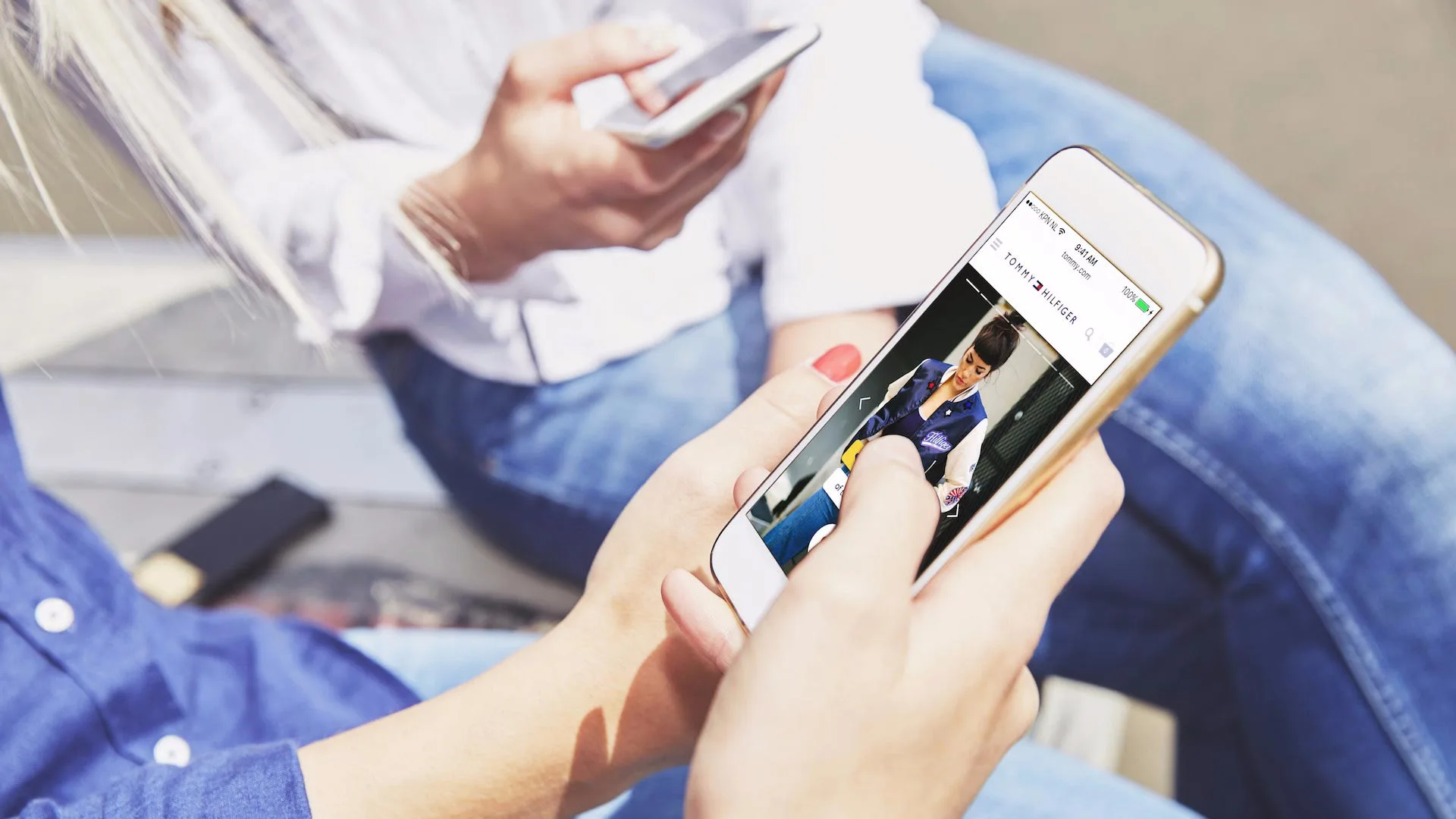 Two people sit close together outdoors, each holding a smartphone. One person is looking at a photo of a child on their phone screen, while the other person is holding their phone in their lap.