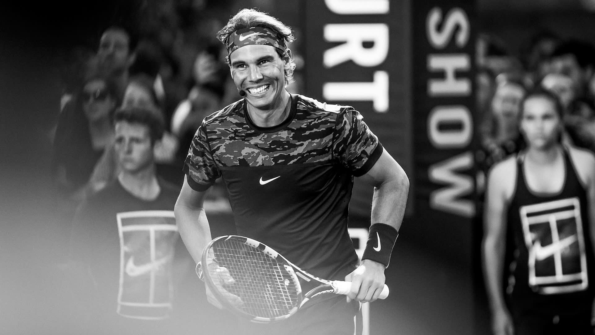 A male tennis player smiling while holding a racket on court, wearing a camouflage-pattern Nike shirt and headband, with people in the background. The image is in black and white.