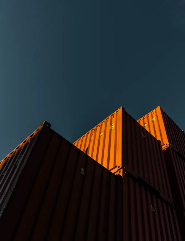 Stacked shipping containers with the top one brightly lit by sunlight against a clear, dark blue sky, creating strong shadows and a dramatic contrast.