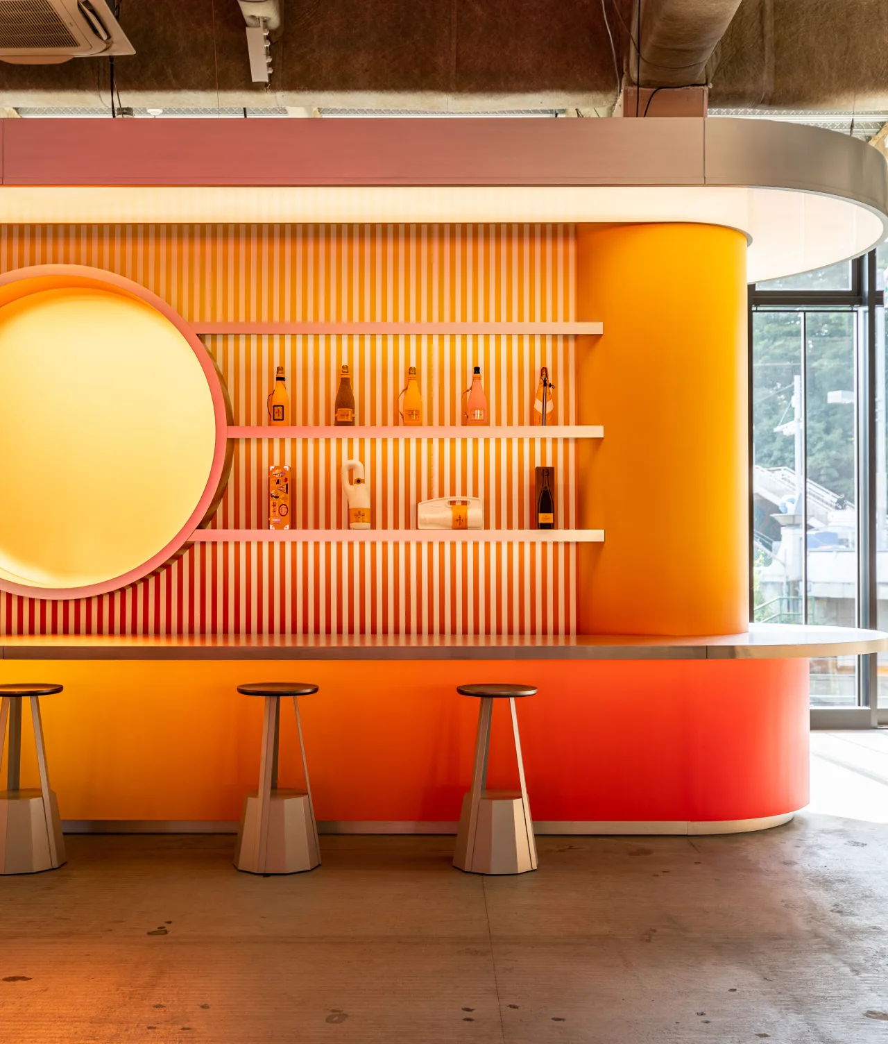 A modern bar with a bright gradient of orange and yellow, shelves displaying liquor bottles, three bar stools, and large windows letting in natural light.