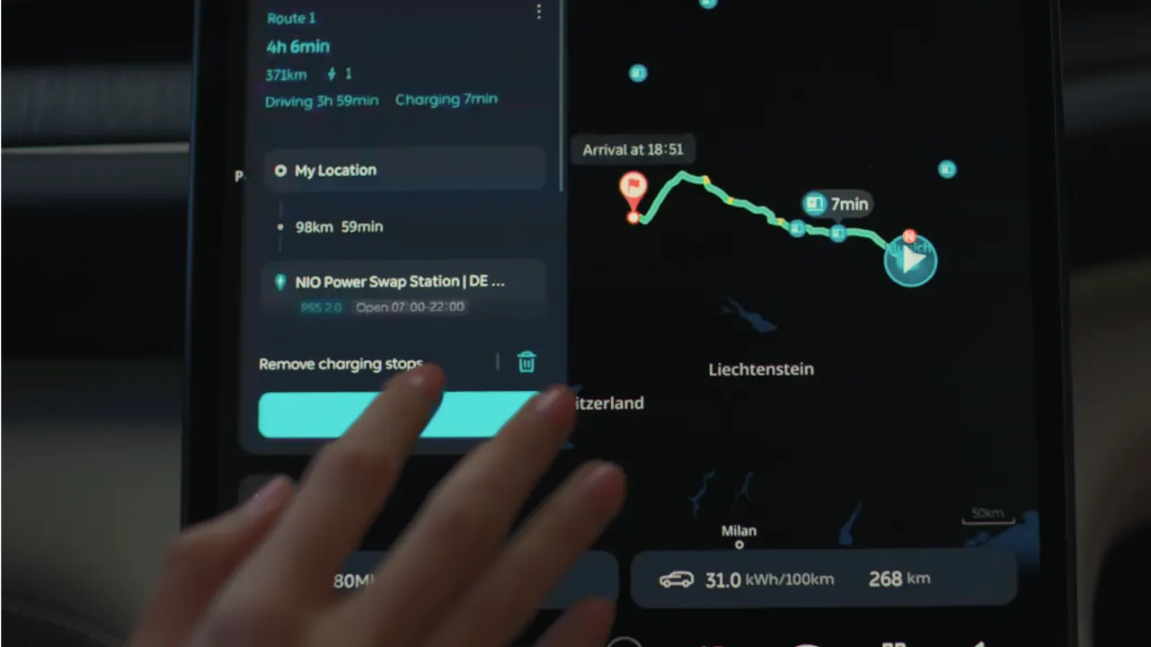A hand interacts with a car navigation touchscreen displaying a mapped route, estimated arrival time, stops, and energy consumption details. The map shows a route passing through Liechtenstein, with Milan visible in the corner.