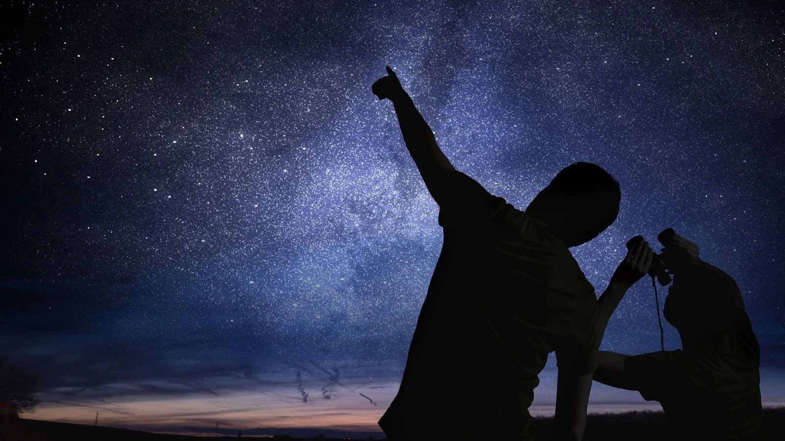 Two people stand beneath a star-filled night sky; one points upward at the stars while the other looks through binoculars, both silhouetted against the glowing Milky Way.