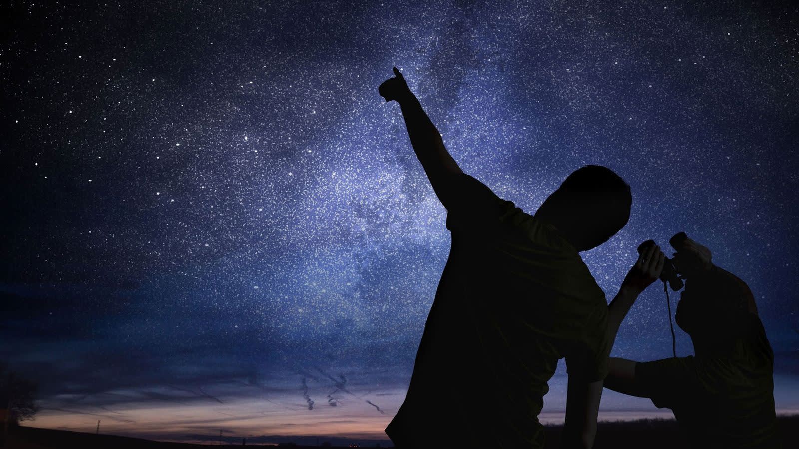 Two people stand beneath a star-filled night sky; one points upward at the stars while the other looks through binoculars, both silhouetted against the glowing Milky Way.