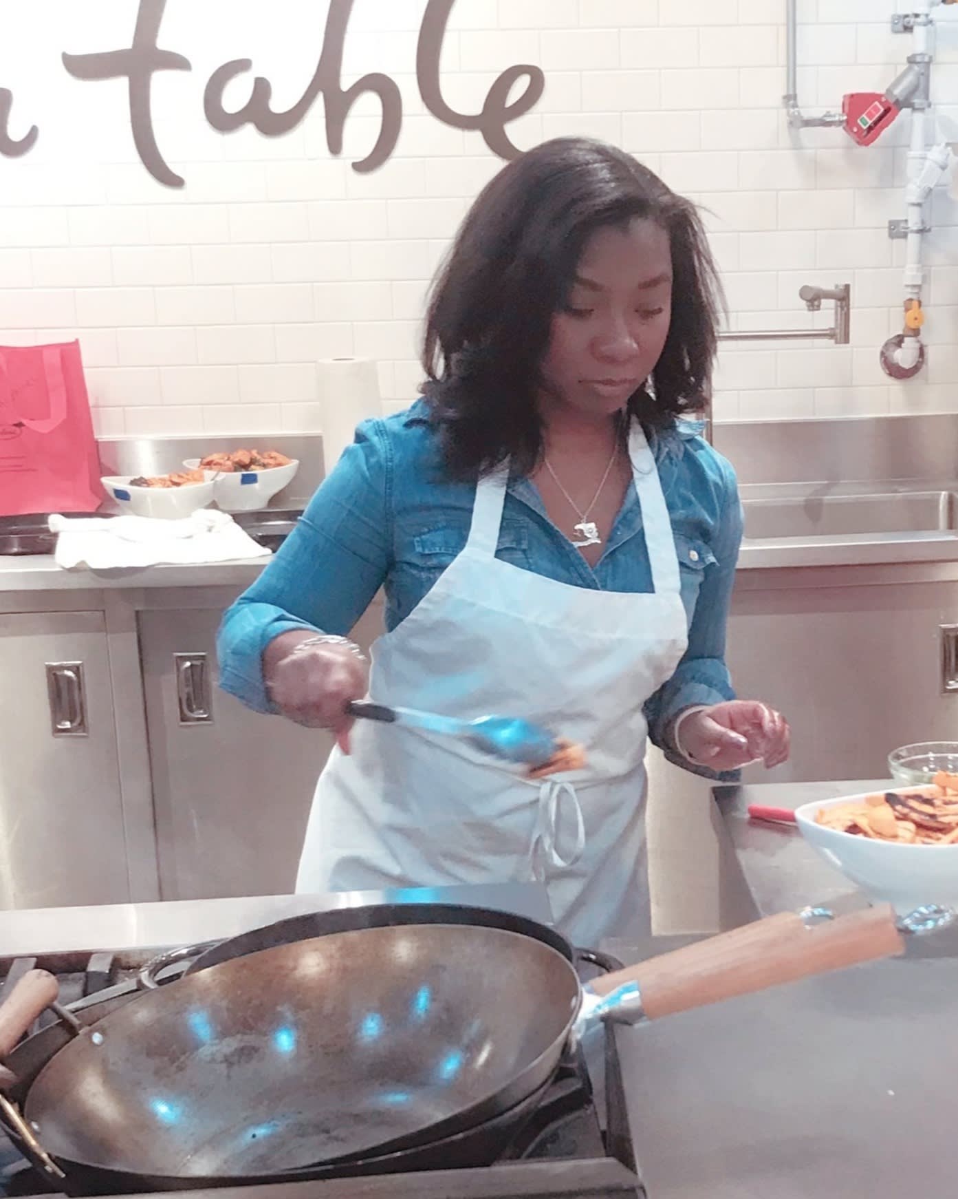 A woman in a denim shirt and white apron cooks in a kitchen, stirring food in a large pan on the stove. A bowl of ingredients and utensils are on the counter beside her. The background has a tiled wall and kitchen supplies.