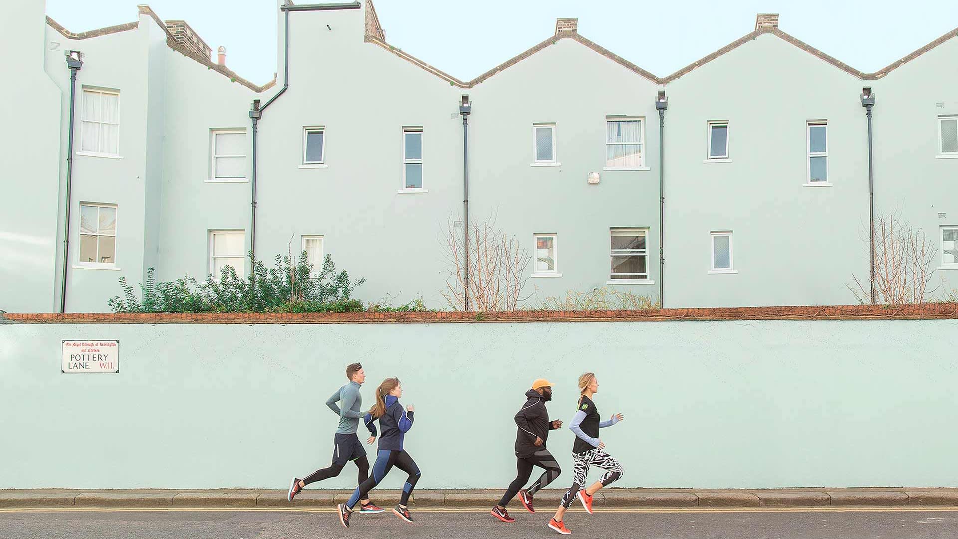 Four people wearing athletic clothing are jogging on a street in front of light blue houses with white-framed windows and a brick wall. A sign on the wall reads POTTERY LANE. The sky is clear.