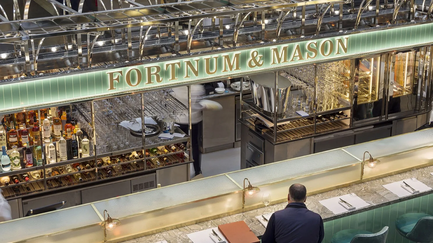 A man sits at a marble counter in front of the Fortnum & Mason bar, featuring a well-lit display of bottles, glasses, and mirrored shelves in a stylish, upscale environment.