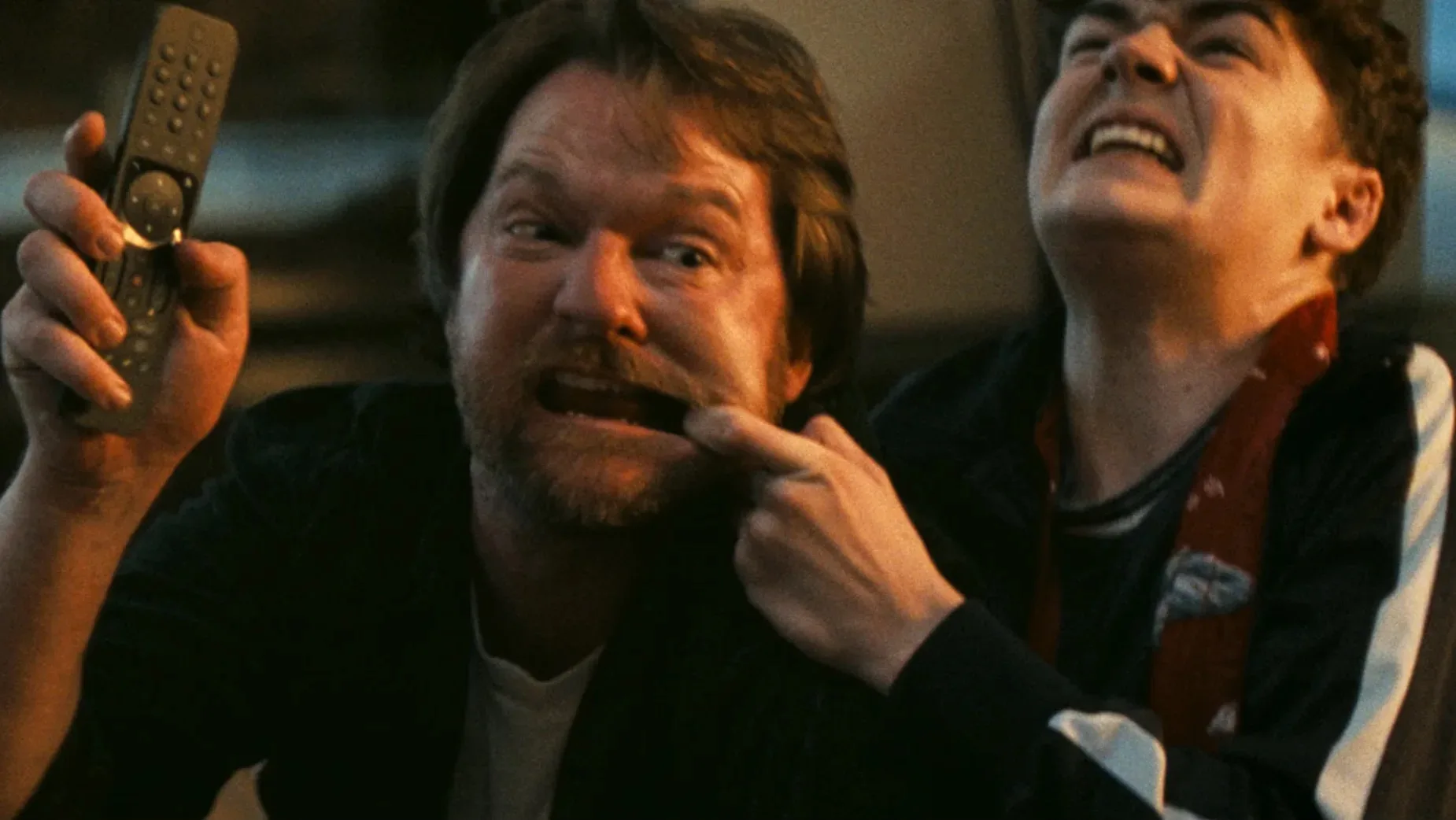 Two people are playfully goofing around; one person is pulling the other’s mustache while holding a remote, both making exaggerated, silly faces. The mood is playful and energetic.