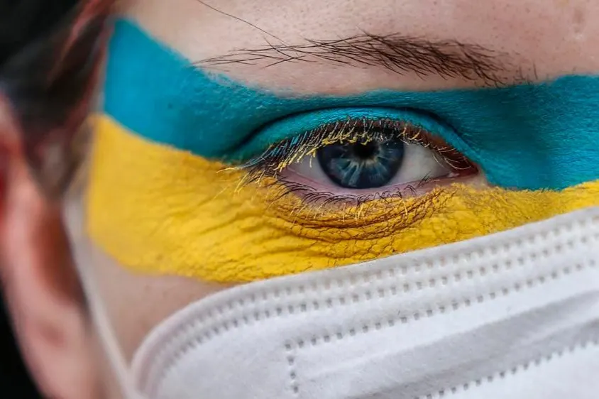 A close-up of a persons face wearing a white mask, with blue and yellow stripes painted across their eye and eyebrow, resembling the Ukrainian flag. The persons eye is visible and looking forward.