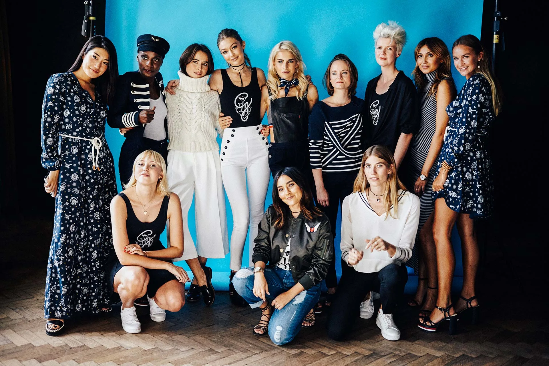 A group of twelve women pose together in front of a blue backdrop, wearing stylish and varied outfits, some standing and some kneeling, smiling and looking at the camera.