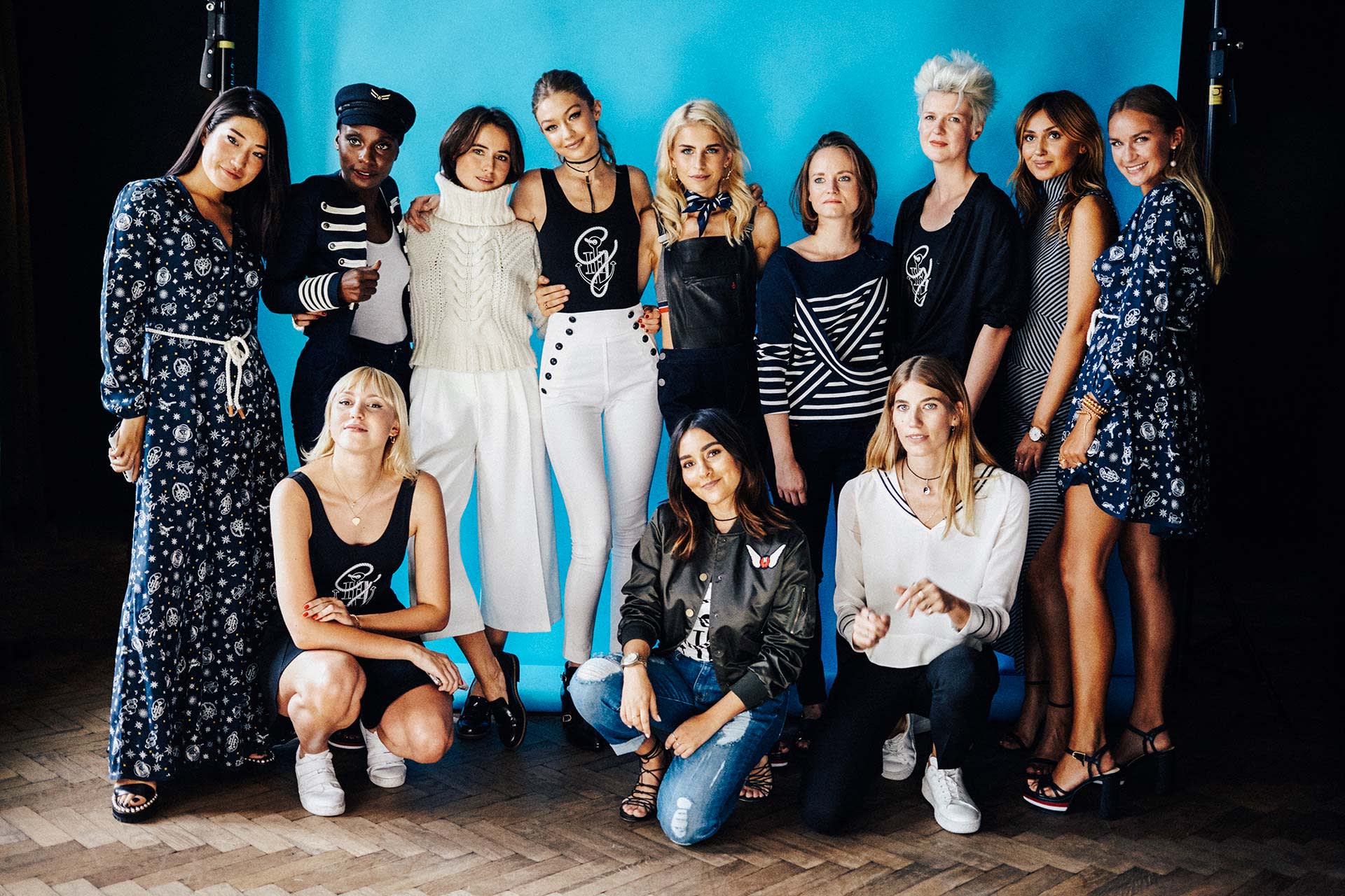A group of twelve women pose together in front of a blue backdrop, wearing stylish and varied outfits, some standing and some kneeling, smiling and looking at the camera.
