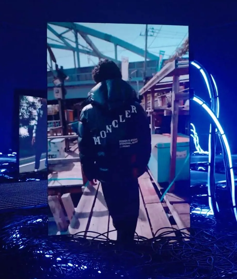 A person wearing a glossy black Moncler jacket walks outdoors along a wooden pathway; the scene is displayed on a vertical screen surrounded by bright blue neon lights and cables.