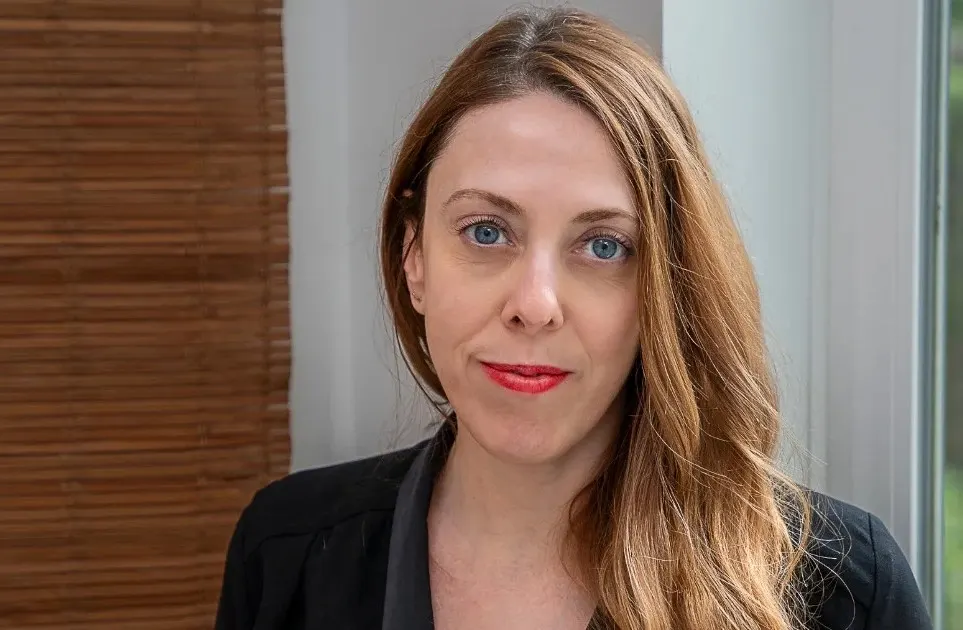 A woman with long light brown hair and blue eyes, wearing a black blazer, stands indoors near a window with bamboo blinds in the background. She has a neutral expression and red lipstick.