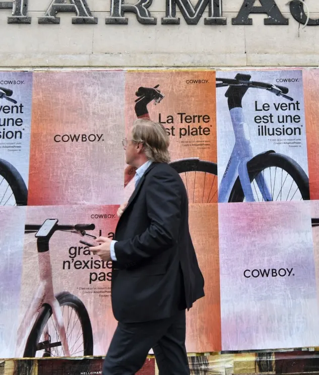 A man in a suit walks past a wall covered with colorful posters featuring bicycle images and French text, including phrases like La Terre nest pas plate and Le vent est une illusion.