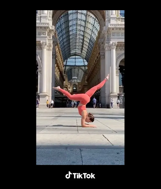 A person in a coral outfit performs a headstand split in an elegant, glass-roofed arcade with ornate architecture, while a few people walk by in the background. TikTok logo is at the bottom.