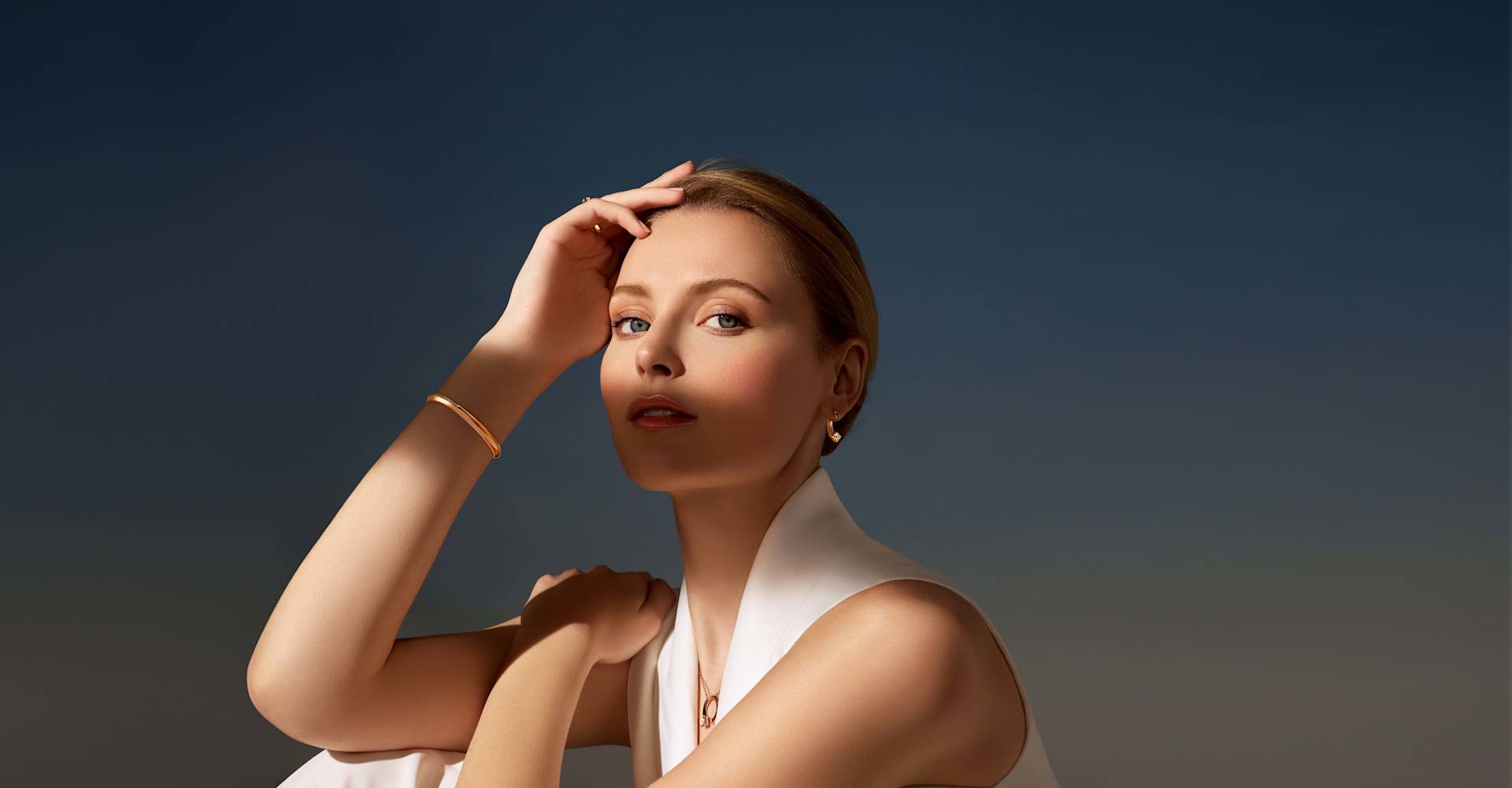 A woman with light skin and blonde hair gazes confidently at the camera, wearing a sleeveless white top and minimal gold jewelry, set against a smooth gradient blue background.