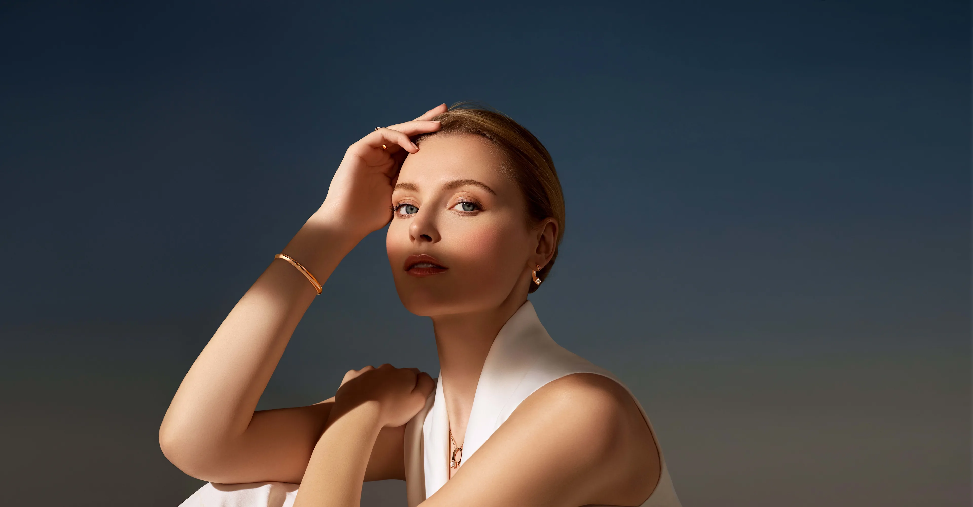 A woman with light skin and blonde hair gazes confidently at the camera, wearing a sleeveless white top and minimal gold jewelry, set against a smooth gradient blue background.